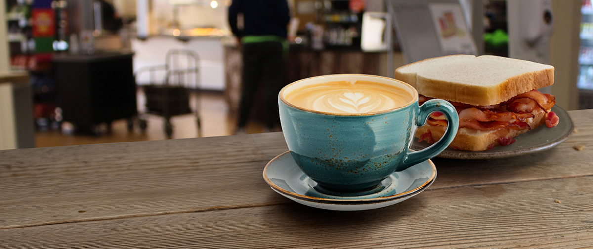 Latte in blue cup and bacon sandwich with tomato sauce on a table in Vanbrugh cafe area